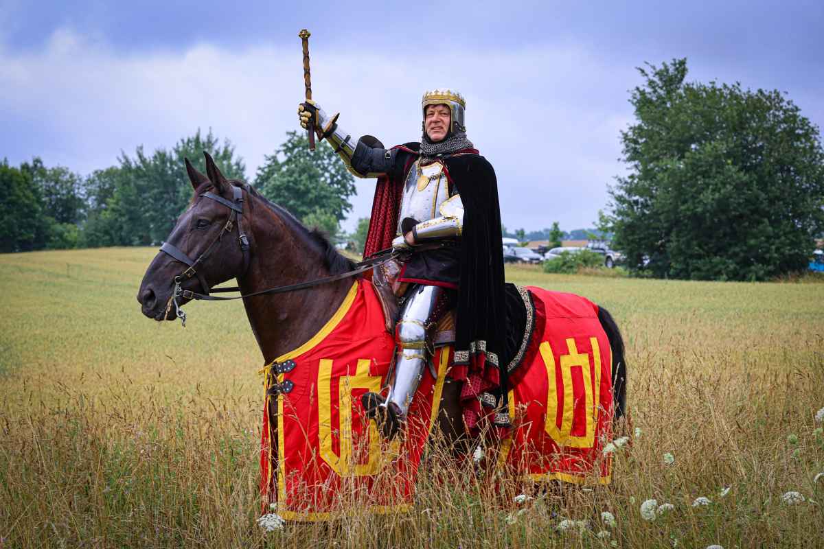 Vyras apsirengęs riterio apranga su šalmu ir kardu sėdi ant žirgo lauko pievoje.